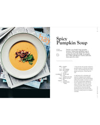 A bowl of creamy pumpkin soup with herbs and seeds sits atop magazines next to Veggies, Every Day of the Week by Cozy Publishing—your go-to inspiration for healthy meals and vegetarian cooking.