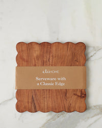 The etúHOME New Scalloped Cutting Board, Medium Square, featuring handmade acacia wood and elegant scalloped edges, rests on a white marble surface with a brown paper “Serveware with a Classic Edge” band.