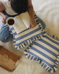 A person sits on a cream surface, holding a cup of tea and reading, with two Casa Amarosa Denim Ruffle 16x16in Pillows nearby and a wooden tray with a coaster.