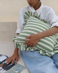 A person in a white eyelet blouse and blue jeans holds the Casa Amarosa Striped Green Ruffle 16x16in Pillow, made from 100% cotton, while resting their hand on an open magazine.