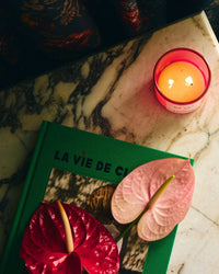 A lit ROEN De Las Flores Candle sits on a marble surface beside a green book titled "La Vie De," with two anthurium flowers inspired by the Mexico City flower market resting gracefully atop the book.