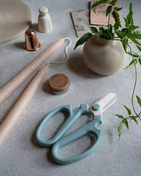 The Floral Society’s Japanese Floral Clippers in Heron, two beige candles, a candle snuffer, a candle helper tin, a white vase with green leaves, and paper items are arranged on a light gray surface.