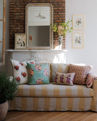 A striped sofa with a Soil to Studio Zeenat Linen Pillow (14 x 20) sits before a white mantle topped with a large mirror, botanical prints, and flowers. In the foreground is a potted plant.