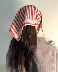 A person with long dark hair, seen from behind, wears Casa Amarosa’s Stripe Hand Block Printed Scarf in Red & Pink and a gray shirt against a softly lit plain background.