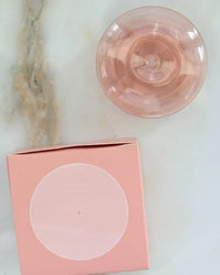 A pink cocktail in a glass sits on white marble next to the BOTANICA Bubble Glass Incense Holder in Coral Pink, a square holder with a circular design that evokes the charm of colored glass.
