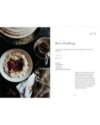 A bowl of rice pudding topped with berries sits on a dark table beside a spoon, textured cloth, cup of tea, and an open copy of "Wonderful Morning - Recipes For A Delicious Start To the Day" by Cozy Publishing.