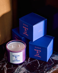 A ROEN Nueve Leones Candle made from a coconut soy wax blend, with double wicks, sits on marble next to three matching purple boxes featuring pink and blue accents and the same branding.