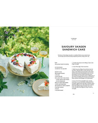 A round savory sandwich cake with white frosting, inspired by Nordic cuisine, is garnished with herbs and flowers beside Cookbook: Crumbs of Joy by Cozy Publishing. A slice is served on a plate next to two glasses of white wine on the outdoor table.