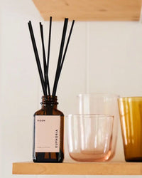 A brown glass ROEN Euphoria Reed Diffuser with black sticks stands on a wooden shelf beside a clear glass and an amber glass, bringing a subtle floral scent to the room.