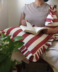A person in a taupe shirt reads while holding two Casa Amarosa Cherry Ruffle 16x16in pillows; the red-and-white striped cotton pillows feature ruffled edges. A green leafy plant is visible in the left foreground.