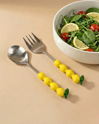 A white bowl with salad—spinach, cherry tomatoes, and lemon slices—sits next to Casa Amarosa’s Salad Servers with Lemon Handles S/2 on a beige surface.