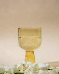 A translucent yellow Aria Glass in Yellow by Casa Amarosa sits filled with a light, bubbly drink on a neutral background, with softly blurred white flowers in the foreground—an elegant piece from their pastel glassware collection.