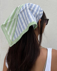 A person with long dark hair, wearing black sunglasses and a white sleeveless top, stands before a plain white wall. They wear the Casa Amarosa Stripe Hand Block Printed Scarf in Green & Blue, facing away from the camera.