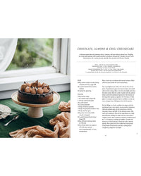 A chocolate almond cheesecake from Cozy Publishing’s "Vegalicious Cheesecakes – Bakery-Style & Plant-Based" rests on a cake stand atop a green tablecloth, next to utensils and the printed vegan cheesecake recipe.