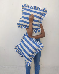 A person in a white tank top and blue jeans holds two Casa Amarosa pillows—one is the Denim Ruffle 16x16in Pillow, covering their face, while the other rests at their side against a plain white wall.