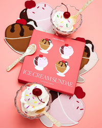Two glass bowls of Neapolitan sundaes with whipped cream, sprinkles, and cherries sit beside a pink Ice Cream Sundae Napkins box by Piecework Puzzles on ice cream-shaped placemats and linen cocktail napkins against a pink background.