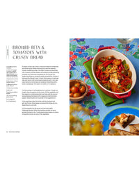A vibrant table displays broiled feta, tomatoes, and olives in a red dish—an ideal self-care meal—from "Snacking Dinners" by Chronicle Books, surrounded by cherry tomatoes, crusty bread, and a blue floral tablecloth.