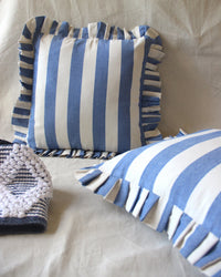 Two Casa Amarosa Denim Ruffle 16x16in pillows with blue and white stripes and ruffled edges rest on a light fabric surface, accompanied by a textured, folded blue-and-white patterned cloth nearby.