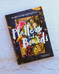 The image features "Platters and Boards" by Chronicle Books, an entertaining cookbook ideal as a hostess gift, on a white wooden surface with a cover showing colorful fruits, cheese, and olives.
