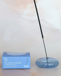A translucent blue BOTANICA Bubble Glass Incense Holder - Blue Moon Glass with a lit incense stick is beside a light blue box labeled "Incense Holder, Handblown Glass, Blue Moon, Translucent" on a neutral background.