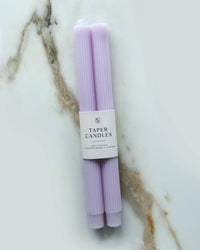 Two unscented, hand-dyed Lavender Taper Candles | Fluted by BOTANICA are bundled with a white Taper Candles label and displayed on a white marble surface with gray and brown veining.