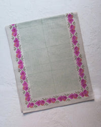 The Simran Table Runner by Soil to Studio features a green center grid and pink floral border on linen, shown flat on a textured white surface. Part of the Valley of Flowers Collection.