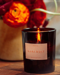 A lit ROEN Ojai Nuit Candle in a dark glass holder rests on a surface, with vibrant orange flowers softly blurred in the background.