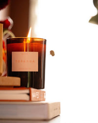 A glowing ROEN Topanga Candle made from coconut soy wax rests on a stack of books, its brown glass softly illuminated against a blurred bright background.