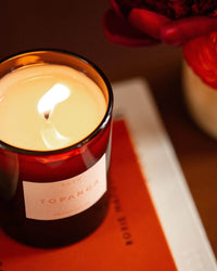 A lit ROEN Topanga Candle, made from coconut soy wax and housed in a dark glass jar, rests on an orange book with a red flower partly visible in the background.