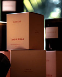 A close-up of stacked ROEN Topanga Candle boxes labeled Made in Small Batch, with glass candle jars behind, featuring our vegan, cruelty-free coconut soy wax Topanga Candle.