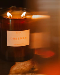 A lit Le Grand Dresden candle by ROEN glows in a dark glass jar, crafted from coconut soy wax with two flames and a warm, blurred background.