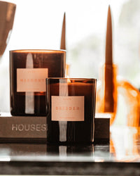Two brown glass ROEN candles—one the vegan soy wax Dresden Candle, the other labeled Marigot—rest on a reflective surface with blurred decor and soft lighting in the background.