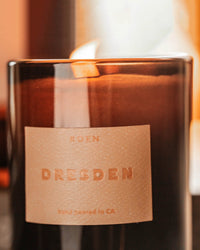 A close-up of the Dresden Candle by ROEN shows ROEN above and hand poured in CA below on a textured beige label. This vegan soy wax candle features an oud blend for a warm, luxurious scent.