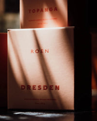 Two candle boxes appear, one labeled Topanga and the other Dresden by ROEN, sunlight casting shadows across their packaging. Each contains a vegan soy wax candle, designed for a clean and eco-friendly burn.