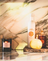 A marble countertop displays a ROEN Ojai Nuit Candle, a white algae cooking oil bottle, a chili oil bottle, and a round yellow object, all illuminated by warm ambient light.