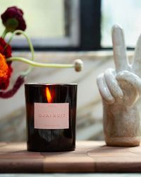 A black ROEN Ojai Nuit Candle sits on a wooden surface beside a white ceramic hand sculpture making a peace sign, with flowers in the background.