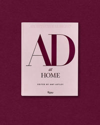 A pink hardcover of "Ad At Home: Architectural Digest" by Random House rests on a matching pink surface, its bold AD letters prominent, offering interior design inspiration.