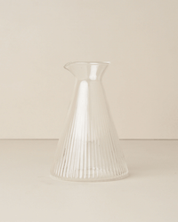 The Saarde Kairos Angular Carafe in Clear, featuring vertical ribbed lines, rests on a neutral surface against a light background, accentuating its elegant design.