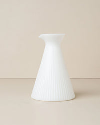 The Kairos Angular Carafe in Opaque White by Saarde features a cone-shaped ceramic form with a narrow spout and subtle vertical ridges, reminiscent of ribbed glass, showcased against a plain beige background.