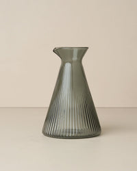 The Saarde Kairos Angular Carafe in Smoke is a ribbed borosilicate glass carafe with a narrow neck and flared base, crafted in translucent gray, shown against a plain beige background.