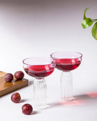 Two Casa Amarosa Cocktail Glasses, handcrafted in India with ribbed stems, are filled with red liquid and placed on a white surface next to four plums, a wooden cutting board, and a green plant hanging from the top right corner.