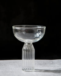 The Casa Amarosa Cocktail Glass, featuring a wide, shallow bowl and ribbed cylindrical stem, is handcrafted in India and displayed on a light-colored surface against a dark background.