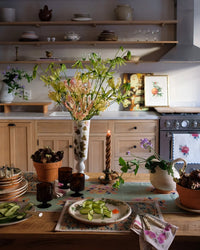 A wooden kitchen table set with cucumber slices, brown glasses, a striped candle, the Dia Table Runner in Sage by Soil to Studio from the Valley of Flowers Collection, potted flowers, and kitchen shelves in the background.