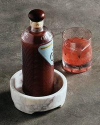 A bottle with a brown cap rests in The Collective’s Cova Wine Coaster, crafted from purple white marble, next to a glass of red beverage on a gray surface.