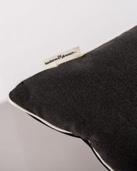 A close-up of the Euro Throw Pillow - Rivie Black by Business & Pleasure Co shows its dark grey color, white edging, and a brand tag. This spill-resistant pillow adds stylish comfort to any outdoor space.