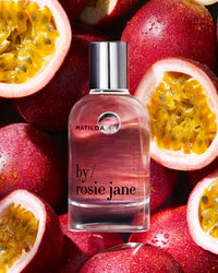 A clear bottle of Rosie Jane Eau De Parfume in Matilda with a silver cap sits atop whole and halved passion fruits, showcasing their vivid yellow pulp and red skins.