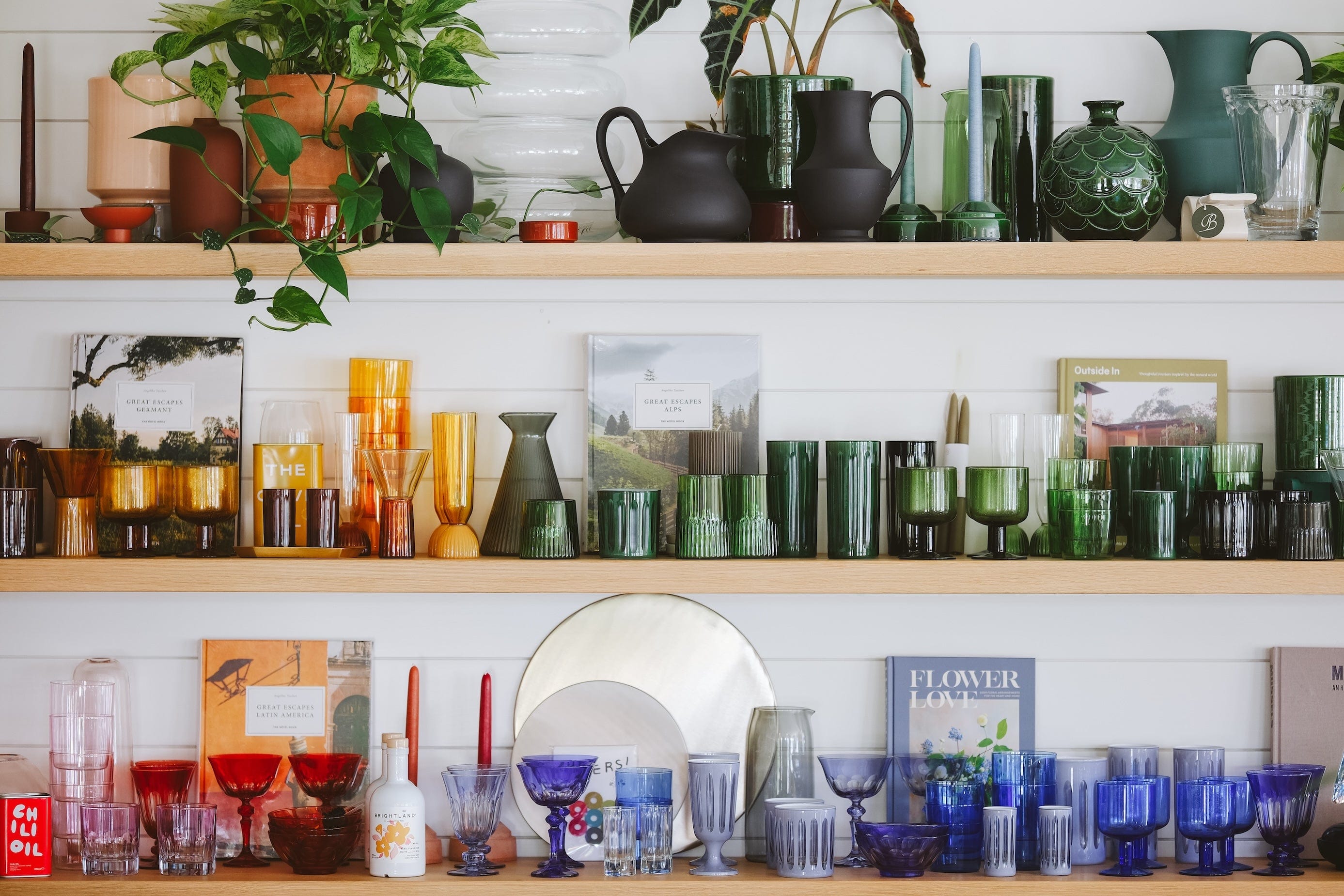 Shelves filled with colorful glassware, books, potted plants, and decorative objects, including vases, cups, pitchers, and bottles in various shapes and colors such as green, blue, red, yellow, and clear glass.
