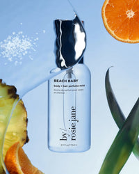 A clear bottle of Rosie Jane Beach Baby Body + Hair Perfume Mist sits among orange and pineapple slices, with sea salt crystals nearby on a light blue background.