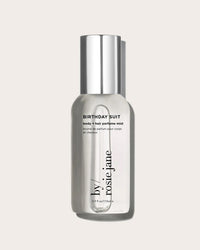 A clear bottle of Rosie Jane Birthday Suit Body + Hair Perfume Mist with a silver cap stands upright on a light beige background. The label displays black text with product details, highlighting this clean, versatile fragrance.
