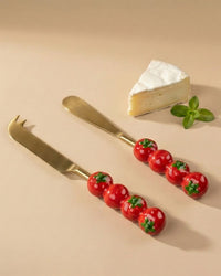 A wedge of cheese, a sprig of mint, and two Casa Amarosa Tomato Cheese Knives S/2 with tomato-shaped handles are artfully arranged on a beige surface.